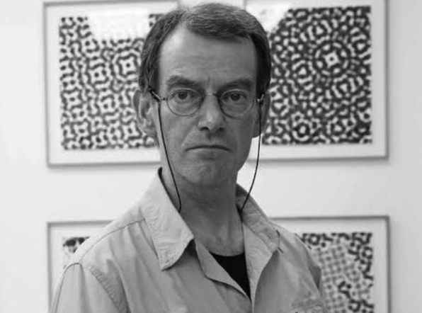 A man with glasses and a serious expression posing in front of patterned artwork in black and white.