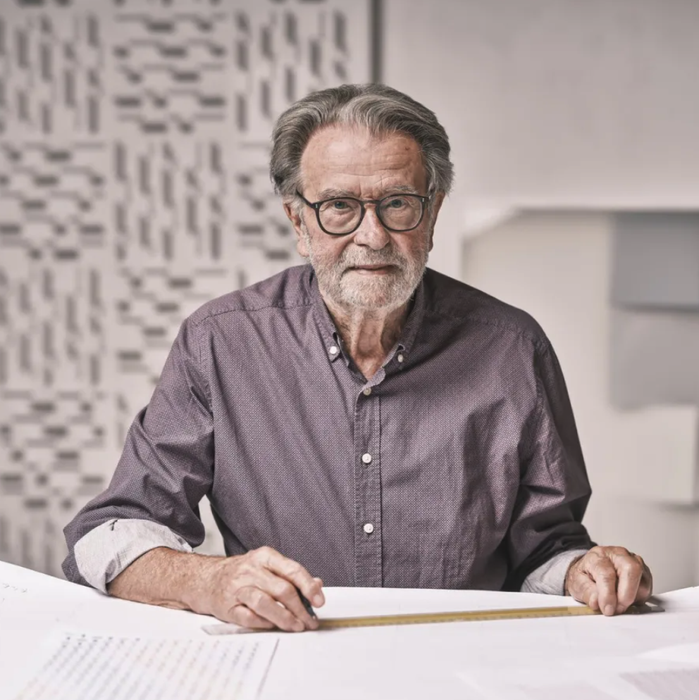 A mature man with glasses and gray hair sitting at a table covered with papers, holding a ruler, with a textured background.