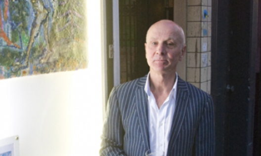 A man in a pinstripe suit stands next to an artwork on a wall, looking slightly serious. The background shows a doorway and part of a building.