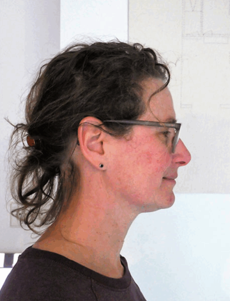 Profile view of a woman with curly hair, wearing glasses, and a dark shirt, standing against a light background.