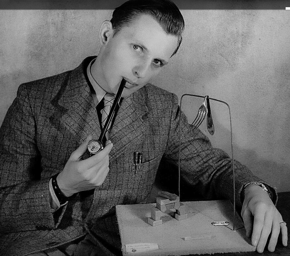 A man in a vintage suit and tie holding a pipe, posing while looking directly at the camera. He is seated next to a model or display with wooden blocks and utensils.
