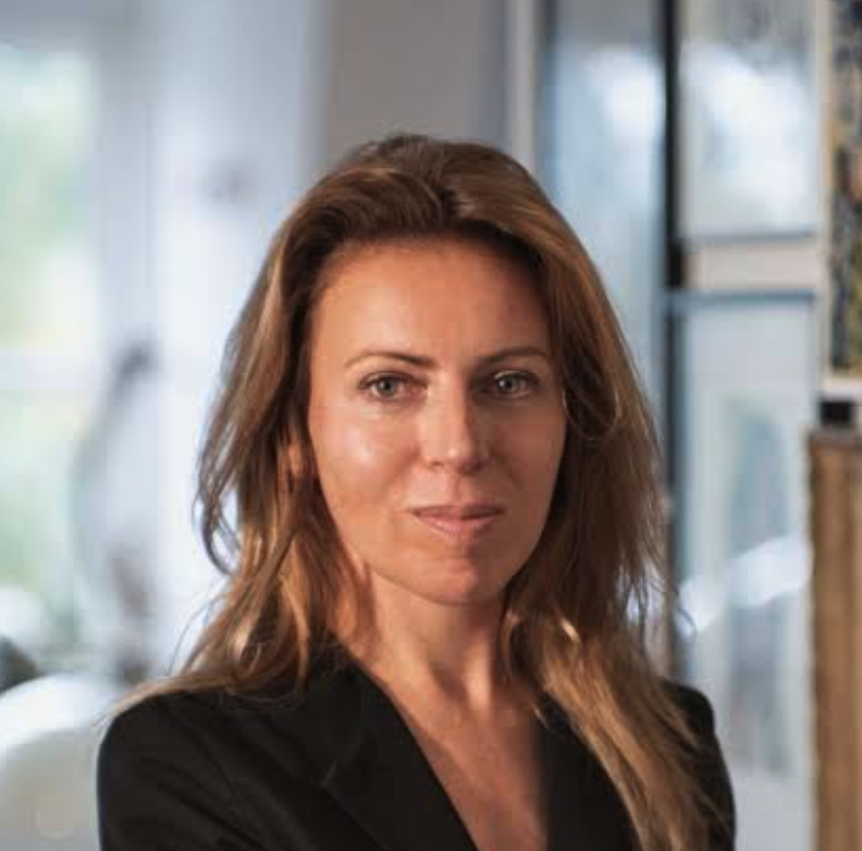 A woman with long, wavy brown hair, wearing a black blazer, stands confidently with a slight smile in an indoor setting filled with soft lighting.