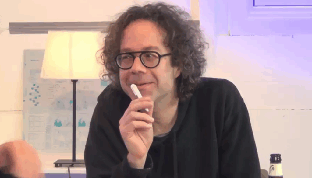 A person with curly hair and glasses sitting at a table, holding a pen and smiling, in a well-lit room with a lamp in the background.