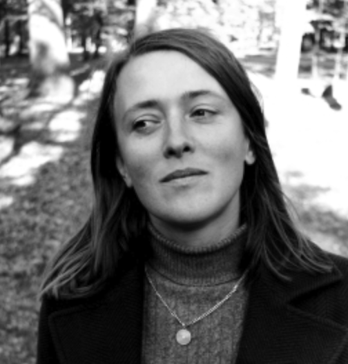 A woman with shoulder-length hair, wearing a turtleneck sweater and a necklace, standing outdoors with a thoughtful expression in a black and white photo.