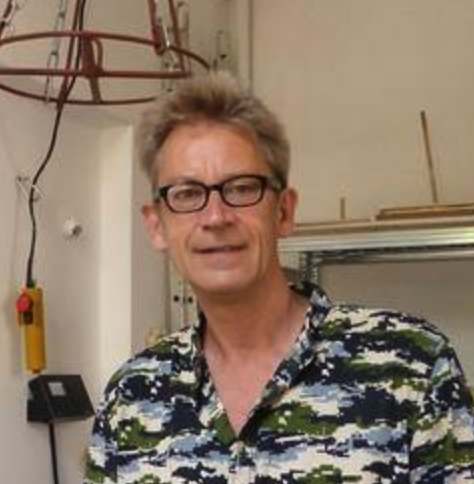 A man with glasses and a patterned shirt, standing in a creative workspace with tools and equipment in the background.