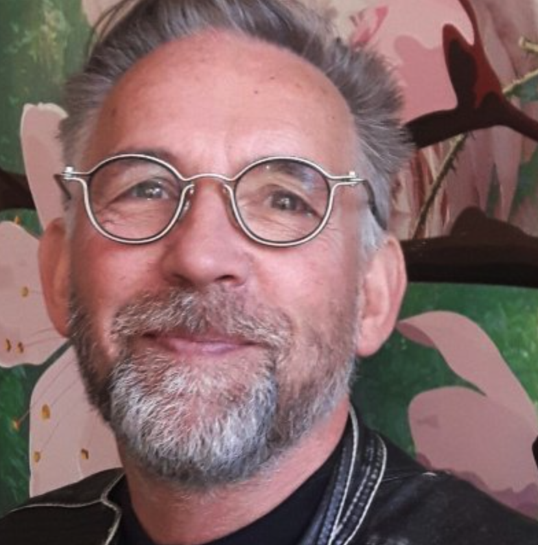 A smiling middle-aged man with glasses and a beard, wearing a black jacket, with a floral background.