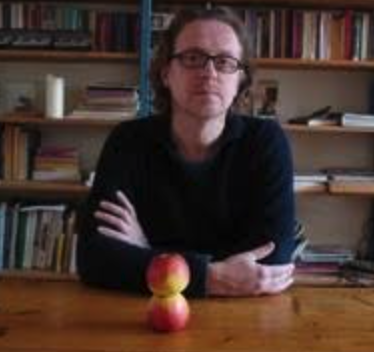 A person with glasses seated at a wooden table with arms crossed, next to two stacked apples and a bookshelf in the background.
