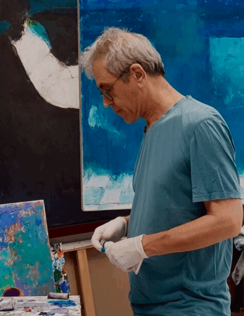 An artist in a teal shirt and white gloves is holding a tube of paint while standing near an easel with abstract blue paintings in the background.