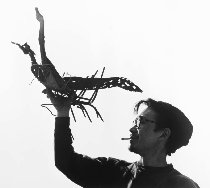 A man holds a handcrafted bird sculpture made from wood and other materials against a bright background, with a cigarette in his mouth and a thoughtful expression.