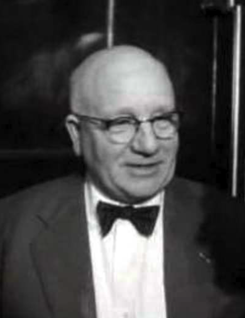 A black and white portrait of a man wearing glasses and a bow tie, smiling and standing in front of a dark background.