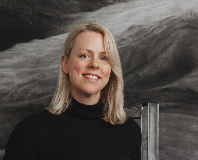 Portrait of a woman with blonde hair, wearing a black turtleneck, smiling against a textured dark background.
