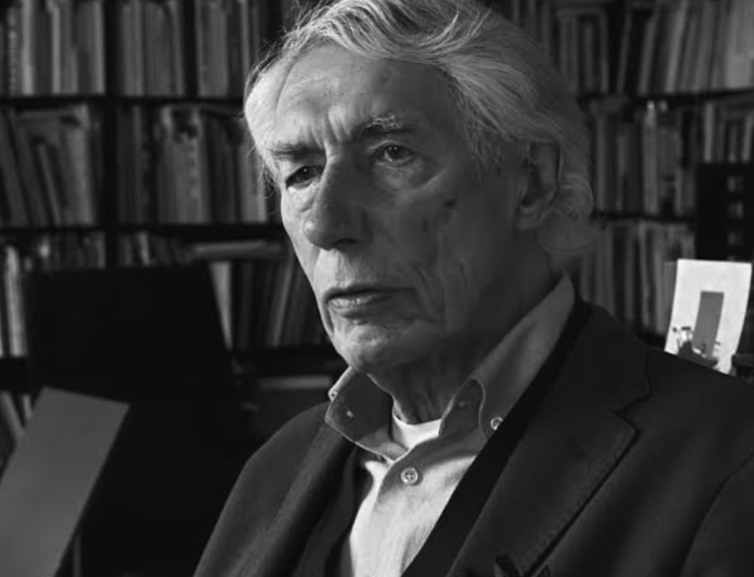 A black and white portrait of a thoughtful older man with grey hair, wearing a blazer and looking off-camera. In the background, there are bookshelves filled with books.