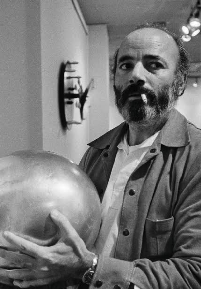 A man with a beard and a cigarette is holding a shiny silver ball while looking at the camera in a gallery setting.