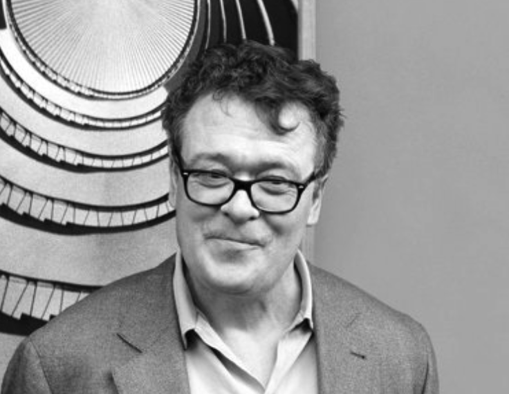 A smiling man with short, curly hair and glasses, wearing a light-colored shirt and a blazer, standing in front of a black and white spiral artwork.