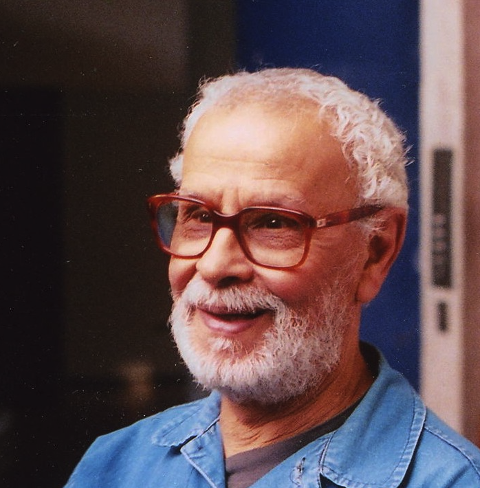 Portrait of an elderly man with glasses and a beard, smiling while wearing a blue shirt.