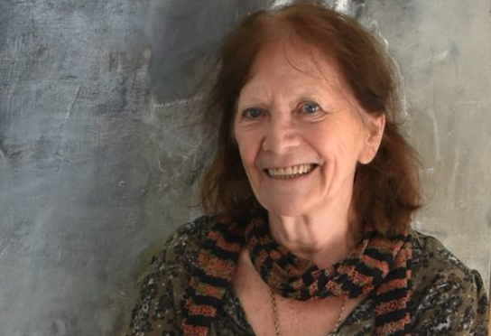 Portrait of an elderly woman with shoulder-length hair, smiling warmly while wearing a patterned scarf against a textured background.