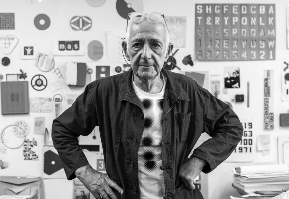 An elderly man in a black and white photo stands with his hands on his hips, wearing glasses and a black jacket, in front of a wall decorated with various colorful design elements and patterns.