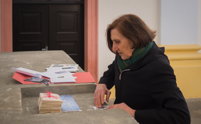 An older woman with dark hair sitting at a table, examining documents and photographs, with various papers scattered around her.