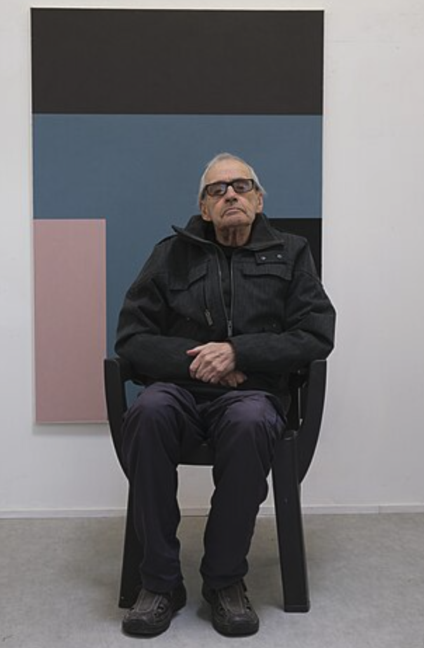 An elderly man sits on a black chair in front of a geometric abstract painting featuring shades of blue, black, and pink.