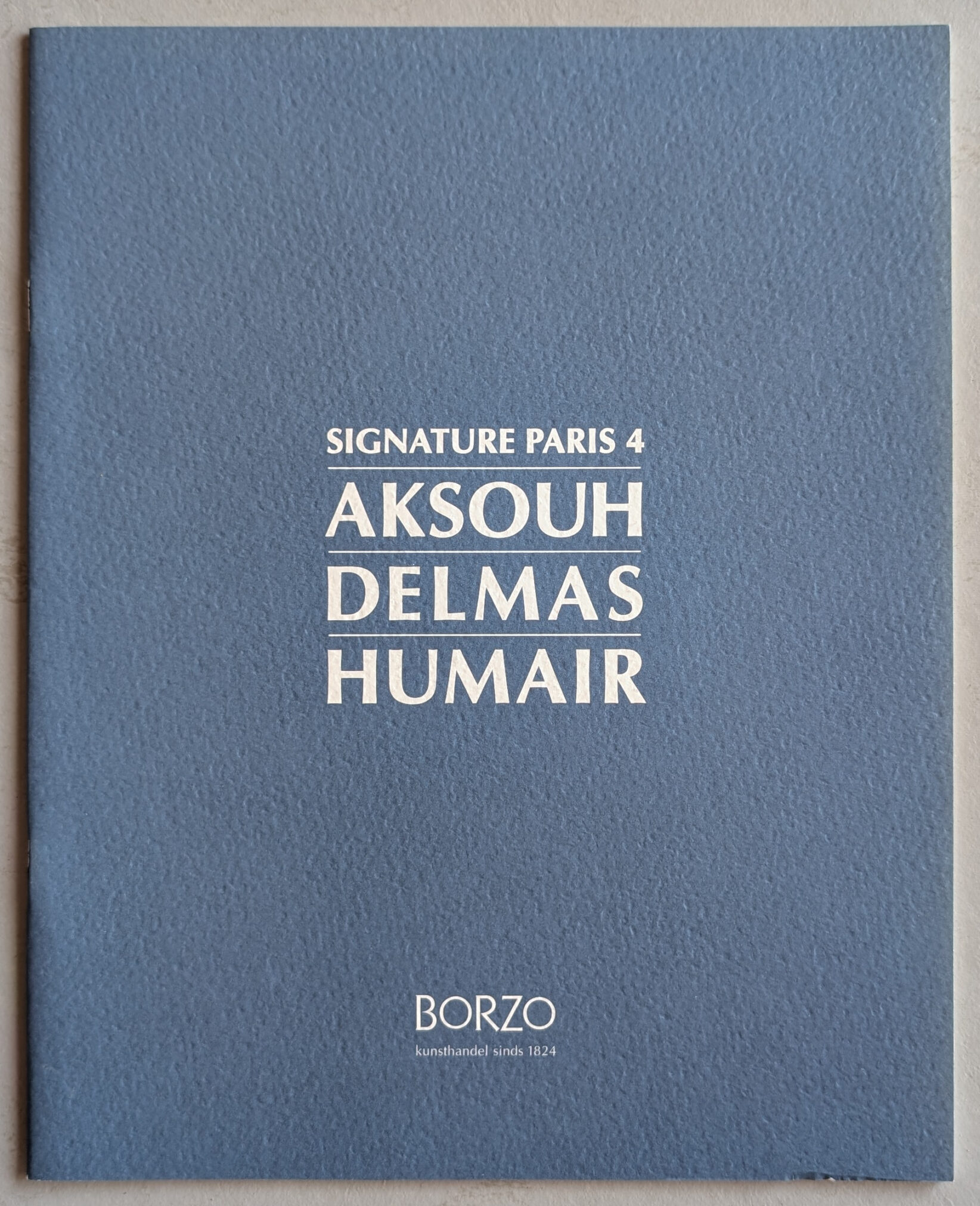 Cover of the catalog for the SIGNATURE PARIS 4 exhibition featuring artists Aksouh, Delmas, and Humair, with a blue background and white text.