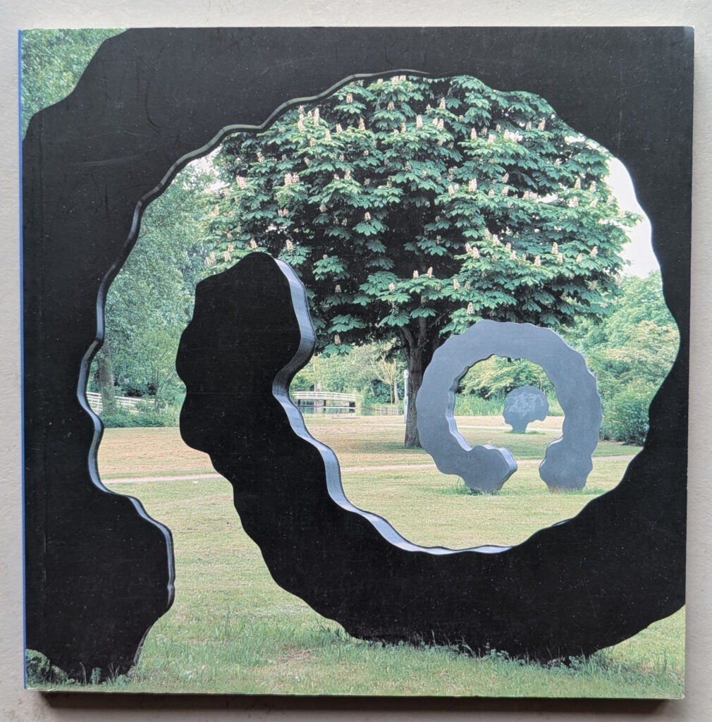 A photograph of a sculptural artwork featuring multiple circular forms in different sizes, set against a backdrop of lush green trees and a grassy area.