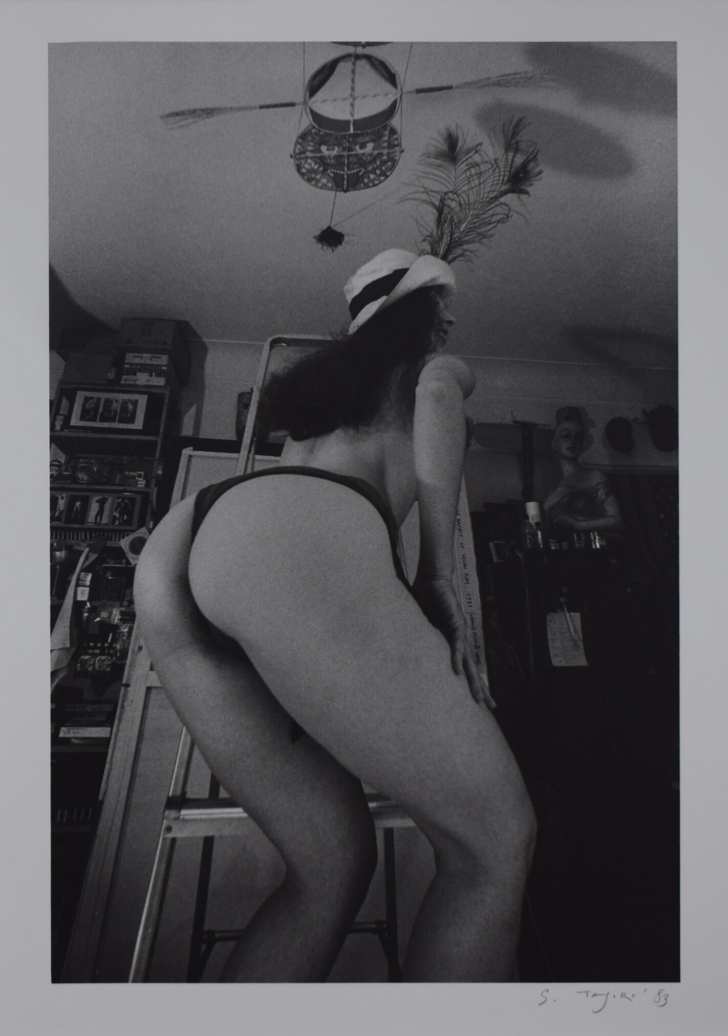 A black and white photograph of a woman posing from behind while standing on a ladder, wearing a hat and minimal clothing in a cluttered indoor setting.