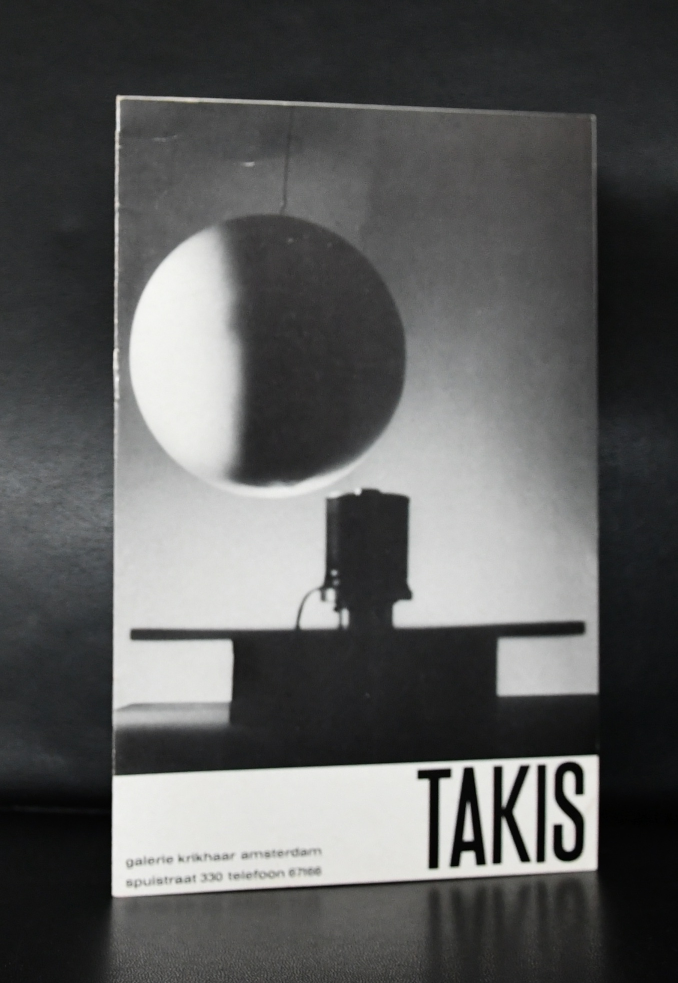 A black and white photograph of a brochure featuring a spherical object suspended above a base, alongside the text 'TAKIS' at the bottom.