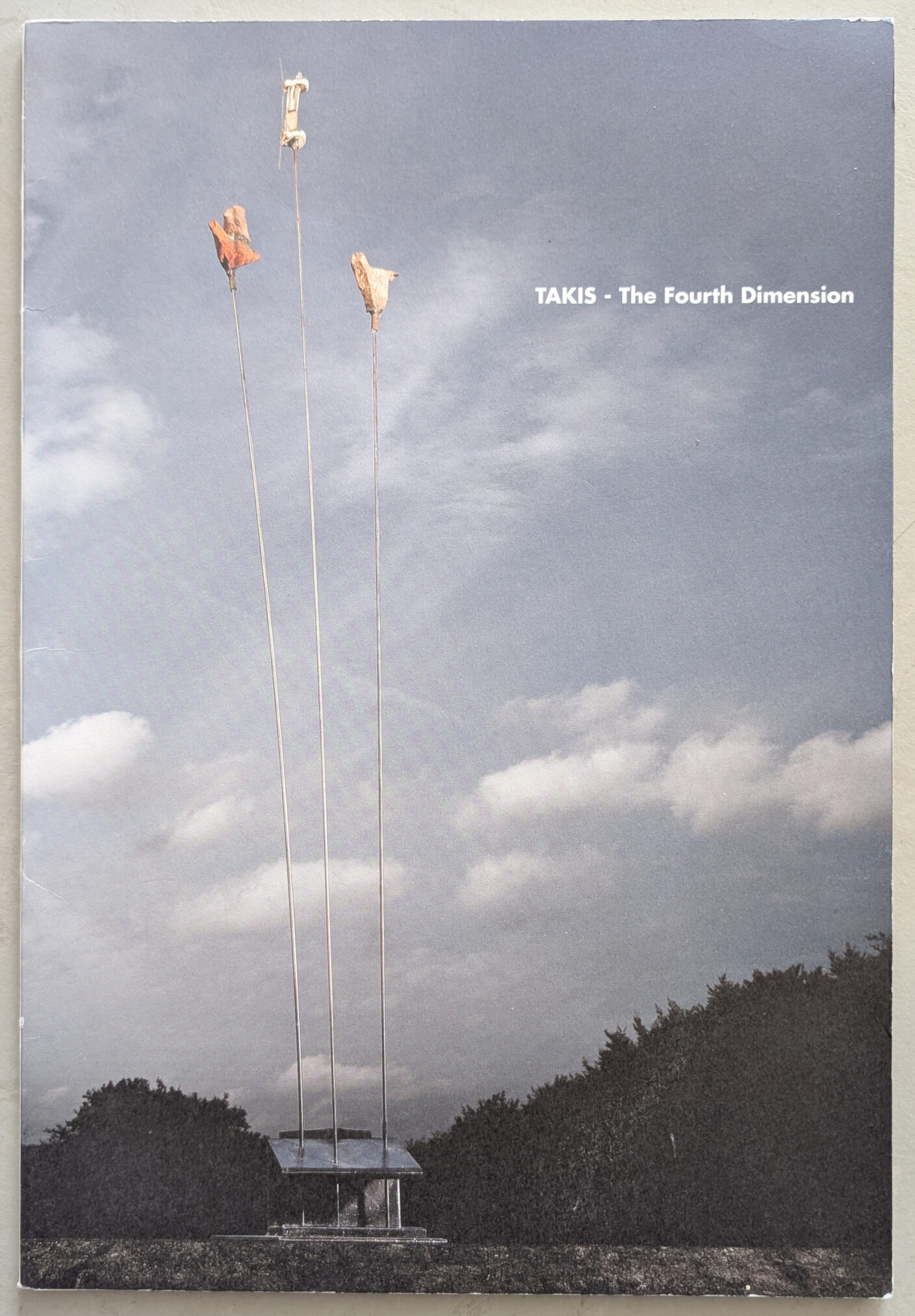 Cover of a publication titled 'TAKIS - The Fourth Dimension', featuring a photograph of an outdoor installation with three tall metal rods, each holding an object at the top, against a cloudy sky.