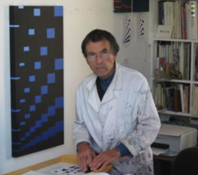 An artist in a white paint-splattered coat stands at a table in a studio, with a geometric black and blue artwork displayed on the wall behind him.
