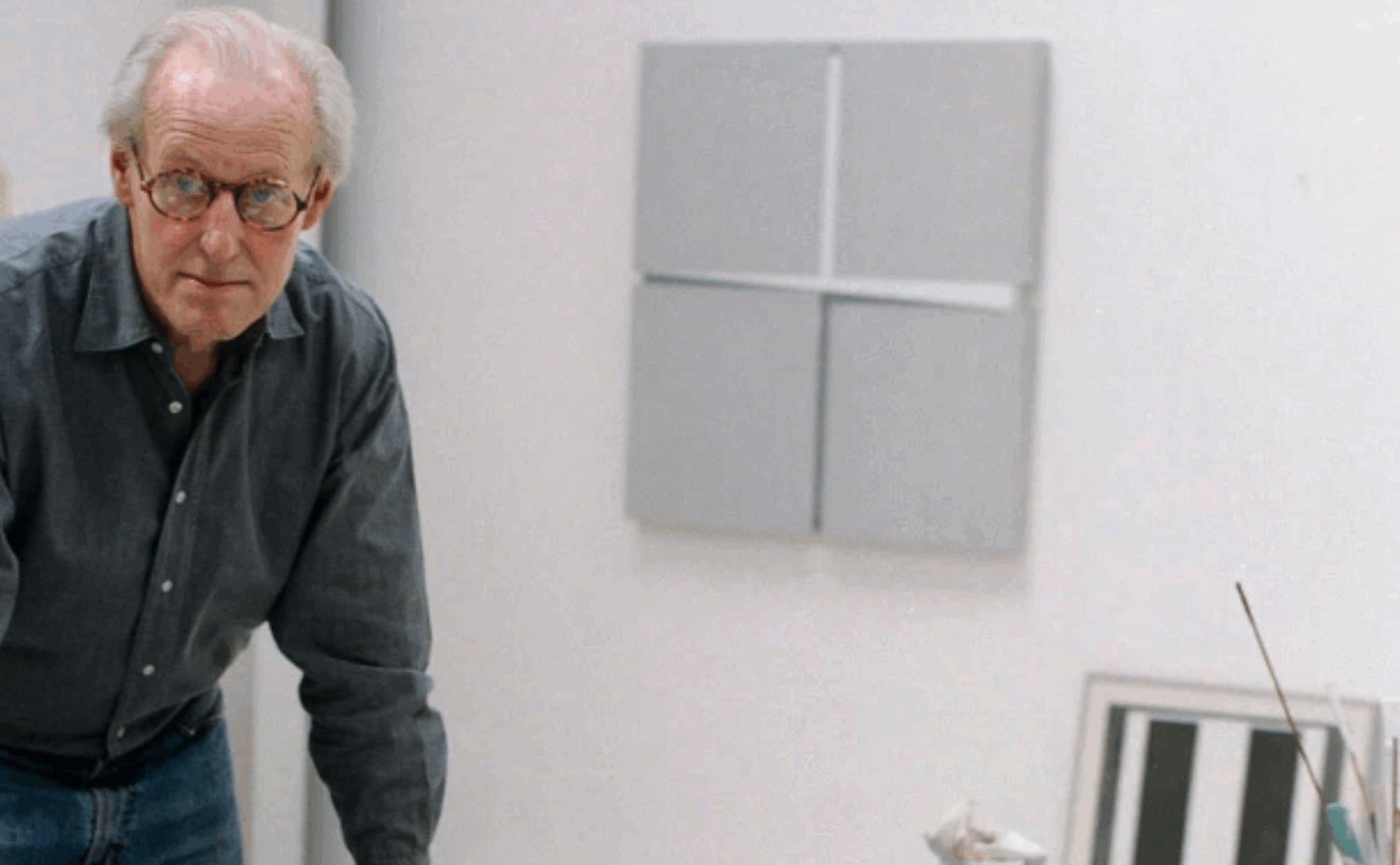 An older man with glasses wearing a blue shirt, standing in an art studio next to abstract artworks on the walls.