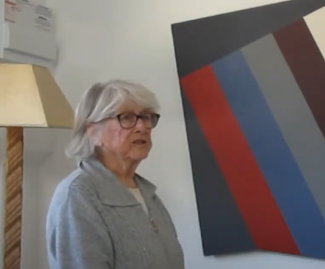 An elderly woman with glasses stands beside a colorful abstract painting, with a lamp in the background.
