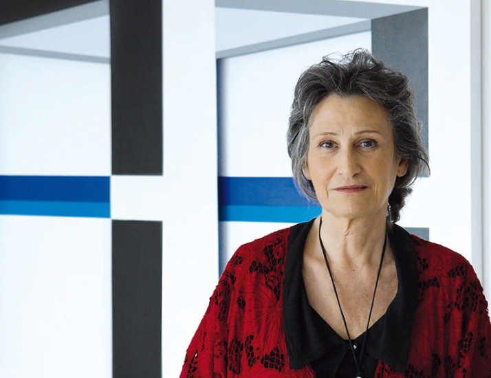 A woman with gray hair wearing a black top and a red patterned cardigan, standing in front of a modern, abstract background.