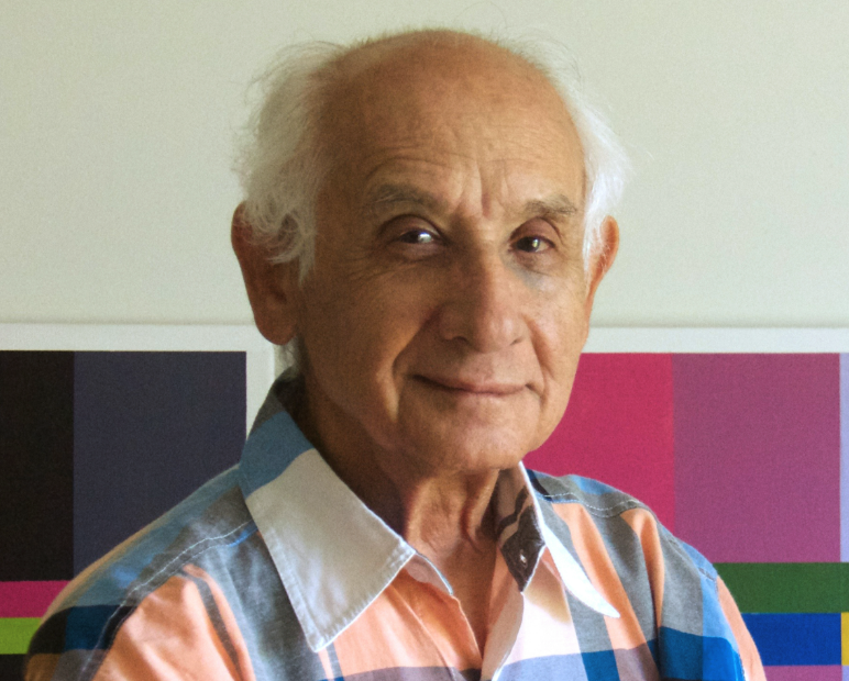A senior man with white hair wearing a colorful plaid shirt, smiling gently in front of a colorful abstract background.