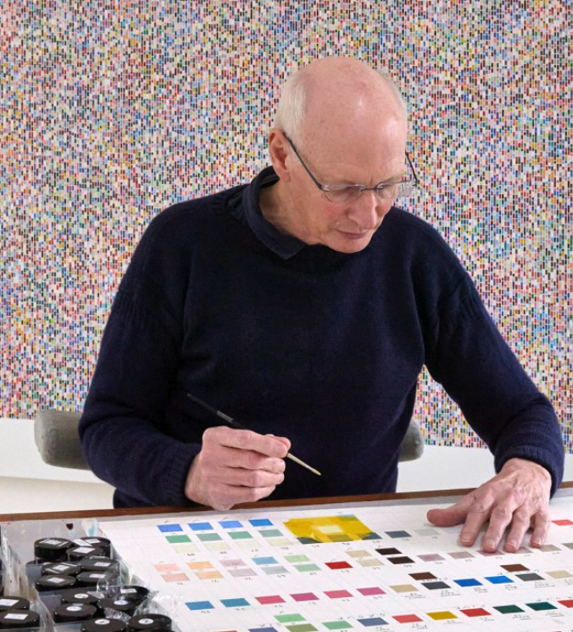 An older man with glasses is sitting at a table, carefully painting colors on a color palette, with a colorful patterned background.