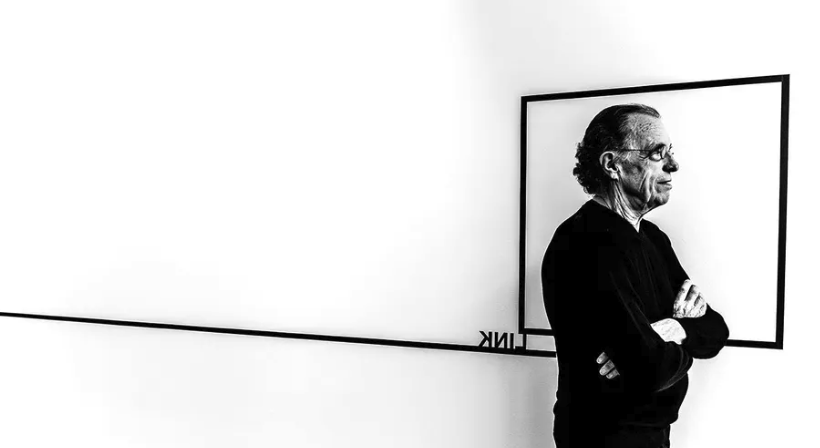 A black and white image of a man standing beside a rectangular frame on a white wall, with a minimalistic geometric line extending from the frame.