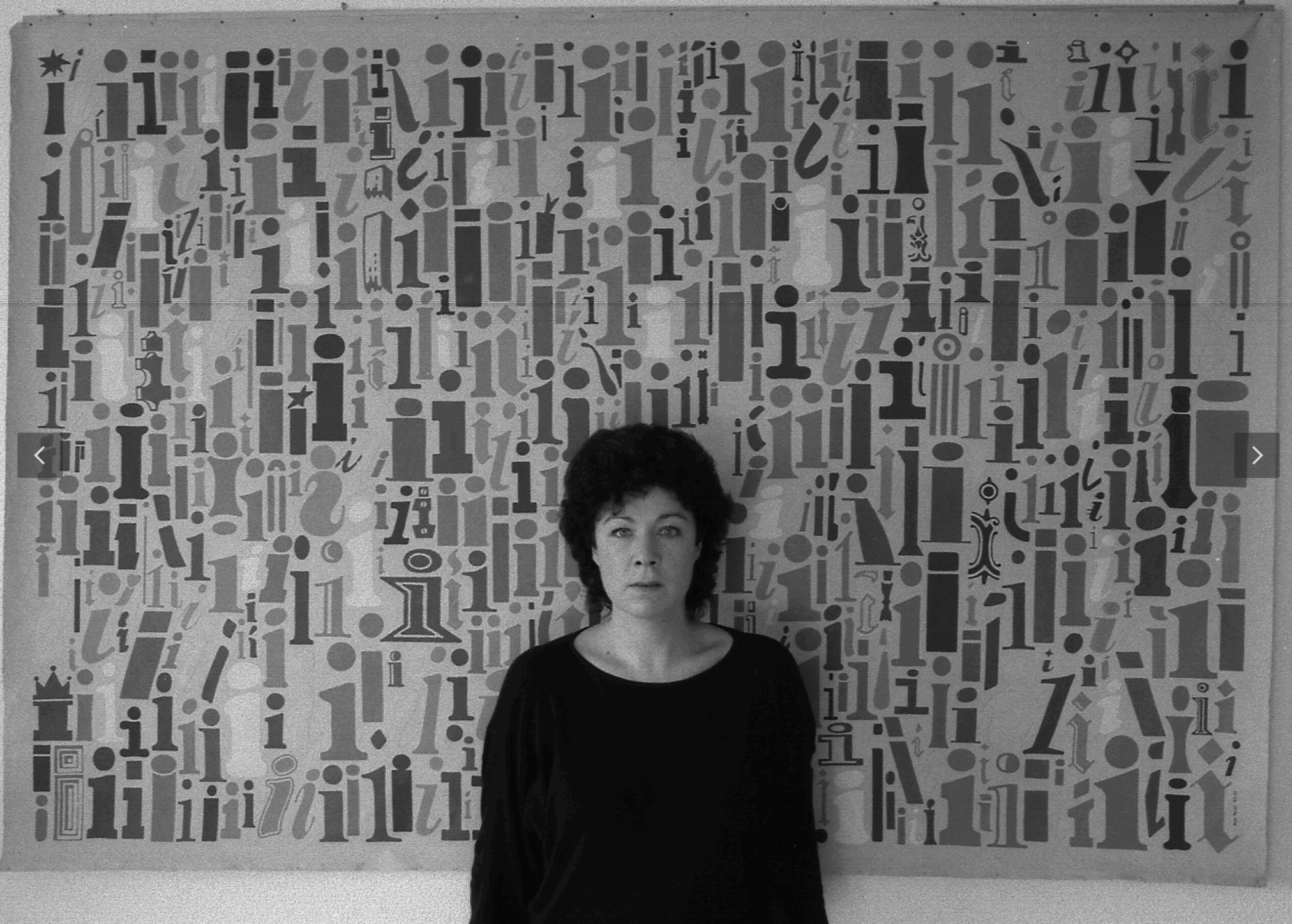 A woman with curly hair stands in front of a colorful abstract wall art featuring various shapes and letters.