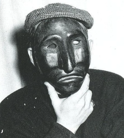 A person wearing a traditional mask and a cap, thoughtfully posing with their hand on their chin.