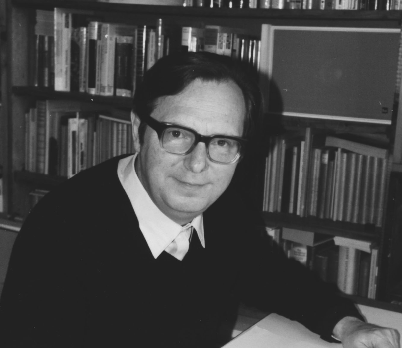 Black and white photo of a man with glasses, sitting at a table in front of a bookshelf filled with books.