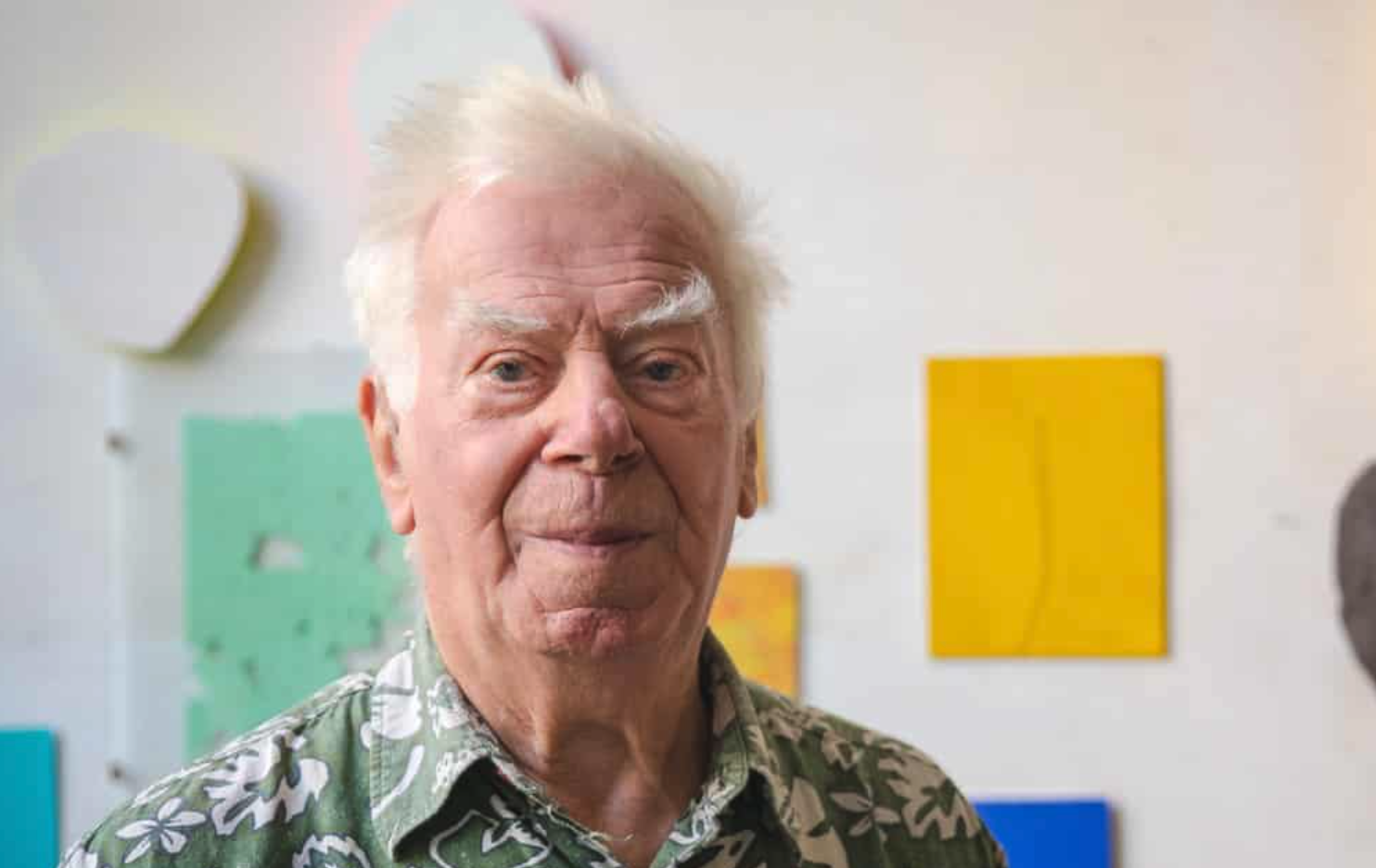 An older man with white hair and a floral shirt smiles in front of colorful abstract art pieces on a wall.