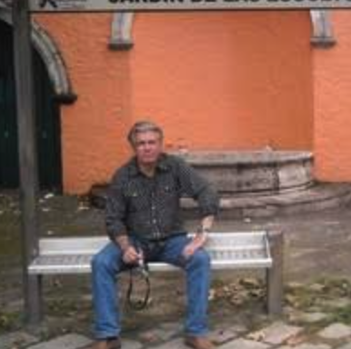 A man sitting on a bench in a park with an orange wall in the background.