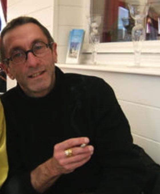 A man with glasses holding a cigarette, sitting indoors with a casual demeanor.