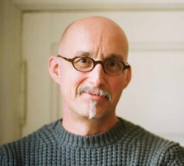 A bald man with glasses and a gray sweater smiles warmly at the camera.