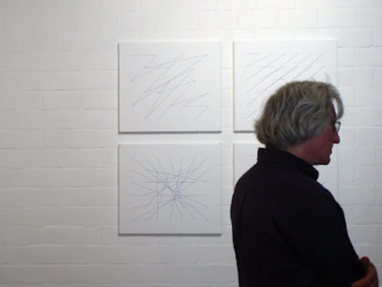 A man with gray hair and glasses is standing next to three abstract line drawings on a white wall.