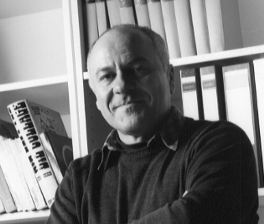 Black and white portrait of a middle-aged man with short hair, smiling slightly, standing with crossed arms in front of a shelf filled with books and files.