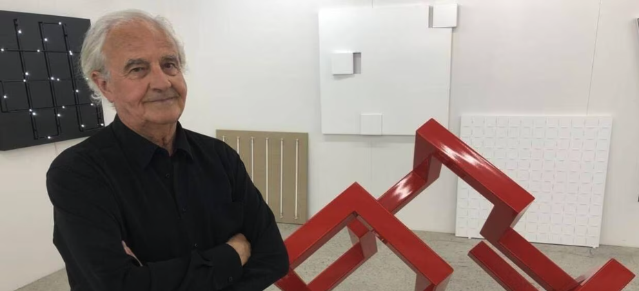 An elderly man in a black shirt stands with arms crossed in an art gallery, featuring modern sculptures and artworks in the background.