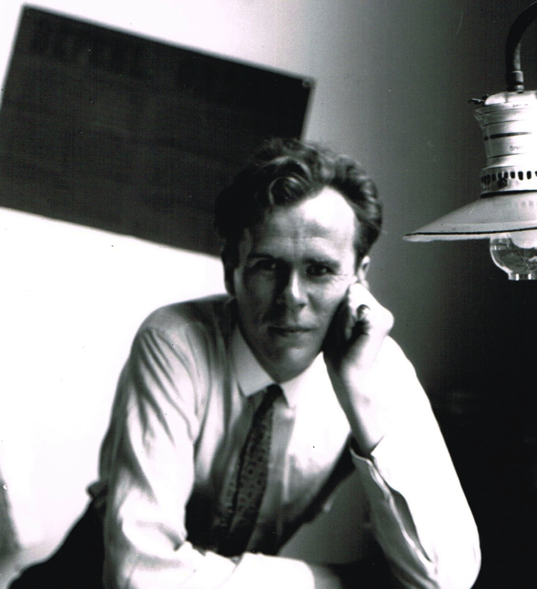 Black and white portrait of a man seated at a desk, resting his chin on his hand, with a lamp hanging beside him.