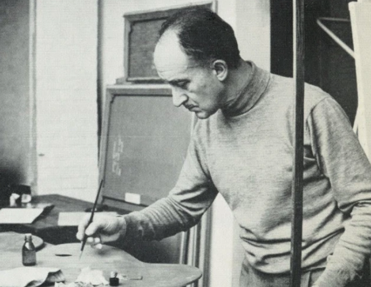 A man in a gray sweater is focused on painting at a table, with various tools and materials in front of him.