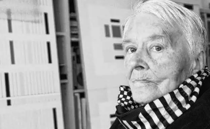 A portrait of an elderly person with short, white hair, wearing a black and white striped scarf, looking thoughtfully at the camera against a backdrop of abstract patterns.