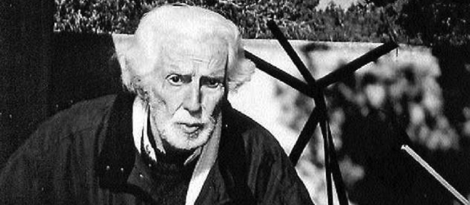 Black and white portrait of an elderly man with white hair and a beard, sitting outdoors with abstract sculptures in the background.