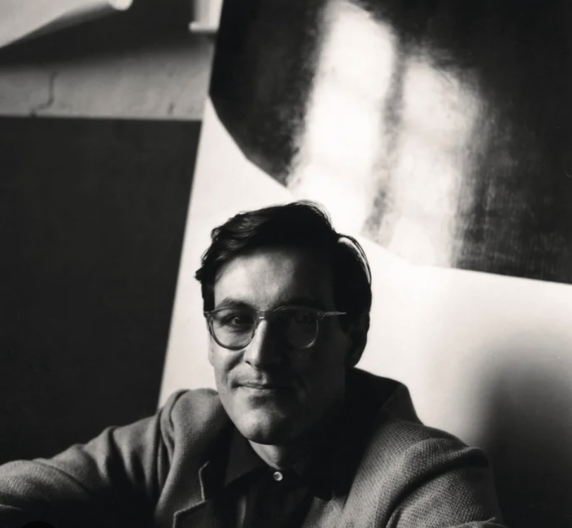 A black and white portrait of a man with glasses, sitting in front of a large abstract artwork.